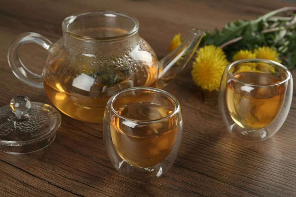 Close-up of herbal dandelion tea in glass teapot and cups on a wooden surface with flowers.