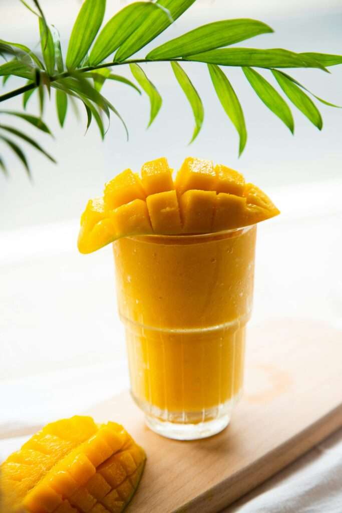 A vibrant mango smoothie in a clear glass topped with fresh sliced mango on a wooden plank.